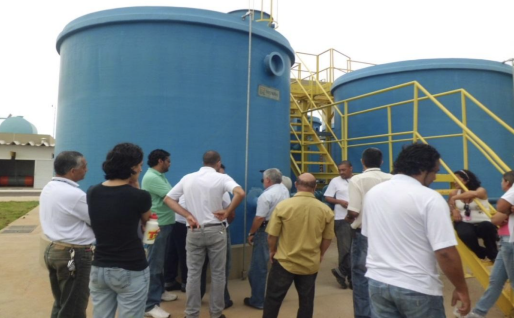 Grupo de profissionais e técnicos realizando visita em estação de tratamento de água com grandes tanques azuis e escadas amarelas.